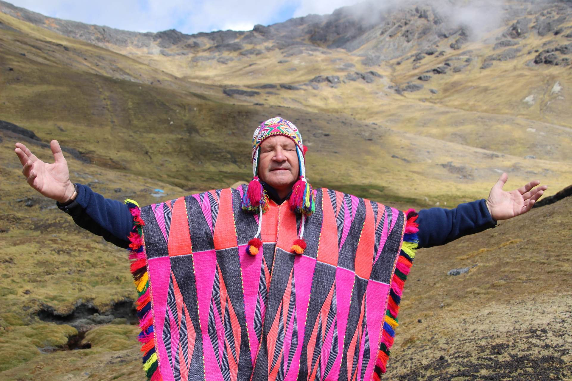 Shamanic practitioner in traditional Q'ero poncho and chullo, arms open in the Peruvian Andes