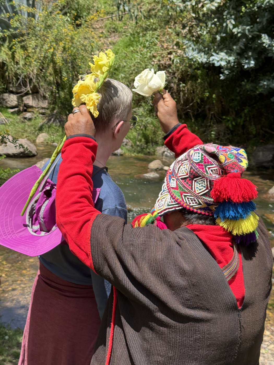 A Q'ero shaman blessing a traveler with flower crowns beside a river